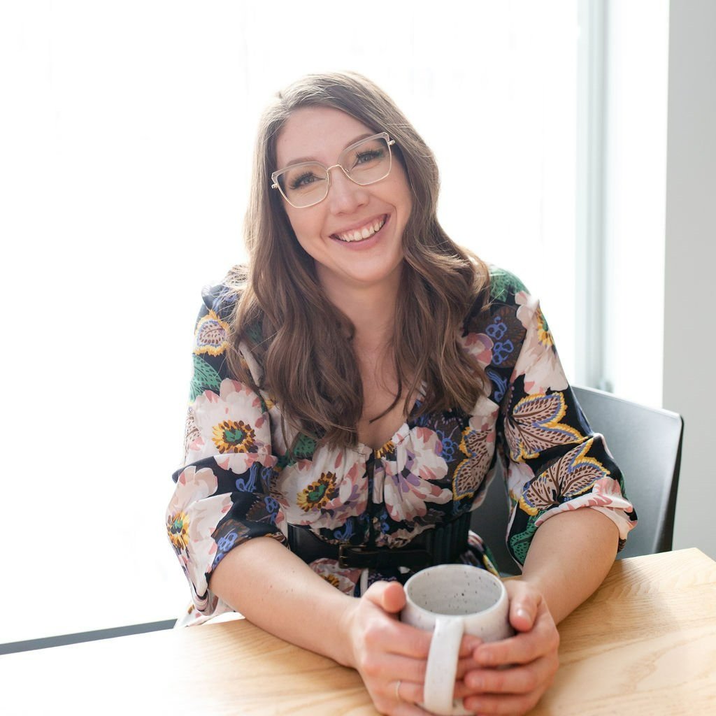 Lindsay smiling at a table with a mug of coffee in her hands.