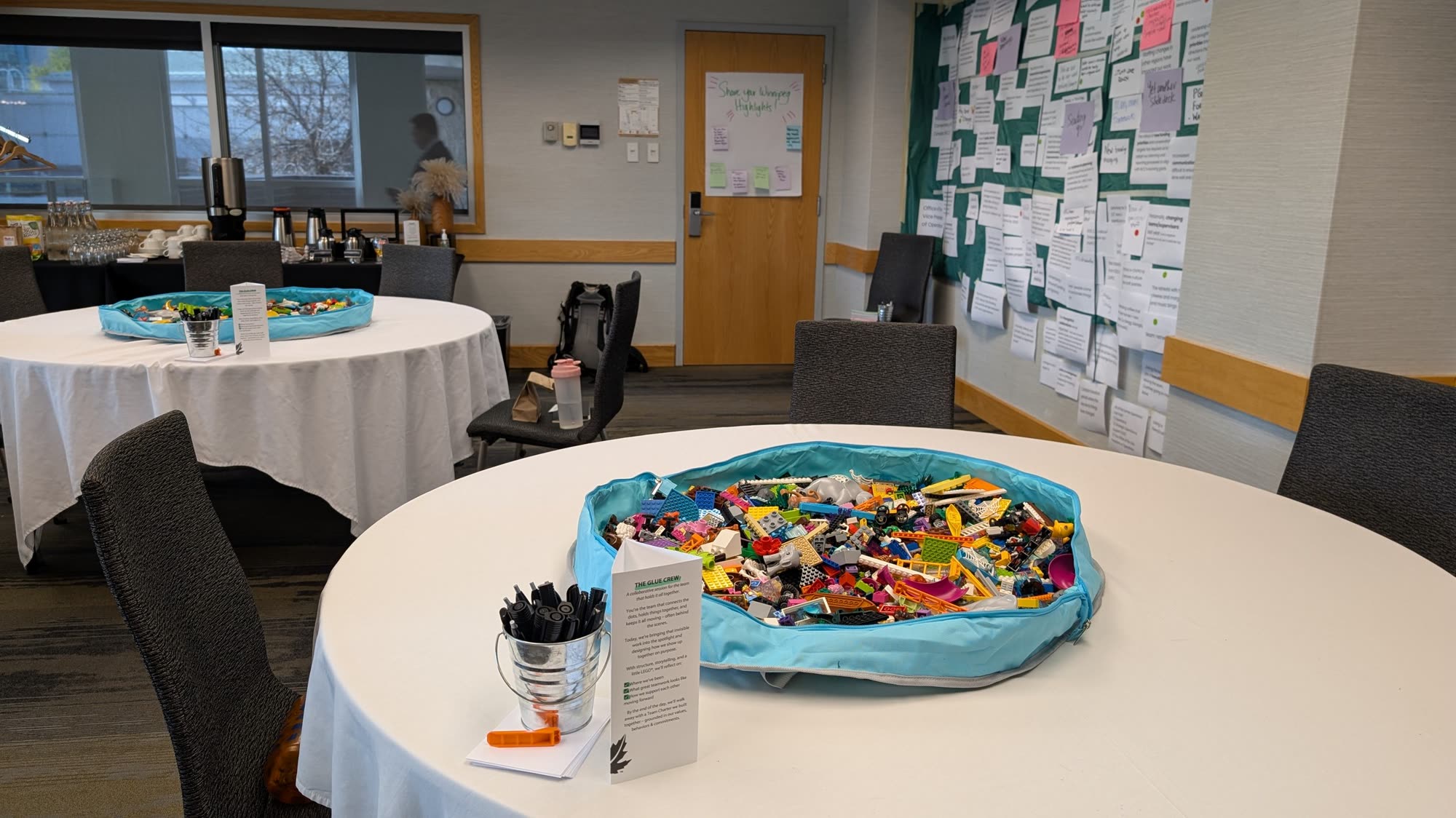 A workshop room set up before people arrive: round tables holding open bins of brightly coloured LEGO and buckets of markers, with a wall behind mapped out in sticky notes.